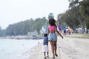 misturado raça africano e ásia mãe e Garoto é jogando às a ao ar livre área. sorridente feliz família ter Diversão corrida em a de praia. retrato do mãe e criança estilo de vida com uma único Penteado. foto