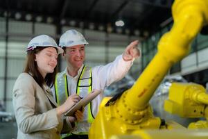 fábrica engenheiros inspecionando em máquina com inteligente tábua. trabalhador trabalho às pesado máquina robô braço. a Soldagem máquina com uma controlo remoto sistema dentro a industrial fábrica. artificial inteligência conceito. foto