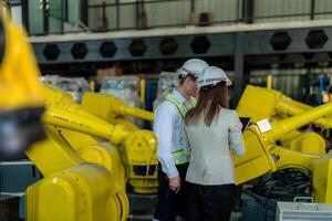 fábrica engenheiros inspecionando em máquina com inteligente tábua. trabalhador trabalho às pesado máquina robô braço. a Soldagem máquina com uma controlo remoto sistema dentro a industrial fábrica. artificial inteligência conceito. foto