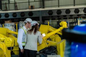 fábrica engenheiros inspecionando em máquina com inteligente tábua. trabalhador trabalho às pesado máquina robô braço. a Soldagem máquina com uma controlo remoto sistema dentro a industrial fábrica. artificial inteligência conceito. foto