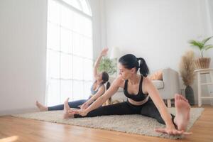 dois mulheres confiante Treinamento ioga. Atlético mulheres dentro roupa de esporte fazendo ginástica alongamento exercícios às casa dentro a vivo sala. esporte e lazer conceito. ioga professor é ajudando jovem mulher. foto