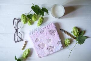 a fechadas Rosa bloco de anotações, caneta, branco vela e galhos do lúpulo Como decoração em uma branco de madeira mesa. Área de Trabalho ainda vida. foto