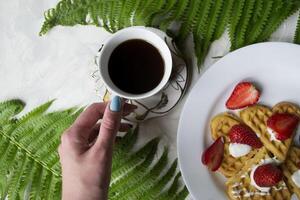 waffles com morango, uma copo do café e verde folhas do samambaia em uma mesa. lindo e saboroso café da manhã. foto