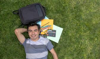 uma jovem aluna homem deitado em a Relva com uma mochila e escola suprimentos foto