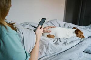 mulher com cachorro arrepiante em cama, usar Smartphone foto