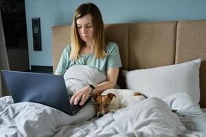 mulher com fofa cachorro deitado dentro cama e usando computador portátil às manhã foto