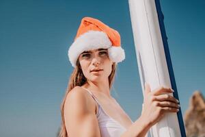 mulher mar e aí. fechar acima retrato do feliz jovem caucasiano mulher com grandes cabelo dentro santa chapéu olhando às Câmera e sorridente. fofa mulher retrato dentro uma branco bikini posando em sup borda dentro a mar foto