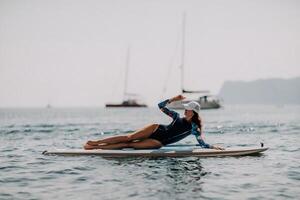 mulher sup ioga. meio era desportivo mulher praticando ioga pilates em remo sup prancha de surfe. fêmea alongamento fazendo exercite-se em mar água. moderno Individual fêmea hipster ao ar livre verão esporte atividade. foto