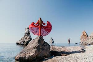 mulher viagem mar. jovem feliz mulher dentro uma grandes vermelho vestir posando em uma de praia perto a mar em fundo do vulcânico rochas, gostar dentro Islândia, partilha viagem aventura viagem foto