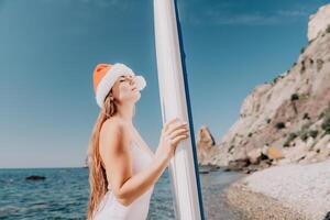 mulher mar e aí. fechar acima retrato do feliz jovem caucasiano mulher com grandes cabelo dentro santa chapéu olhando às Câmera e sorridente. fofa mulher retrato dentro uma branco bikini posando em sup borda dentro a mar foto