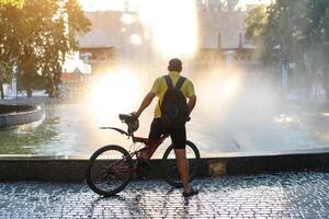 irreconhecível ciclista carrinhos perto fonte segurando bicicleta dentro mãos em uma brilhante ensolarado verão dia. foto