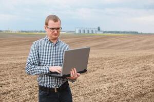 feliz agricultor com computador portátil em pé dentro trigo campo dentro frente do grão silo foto