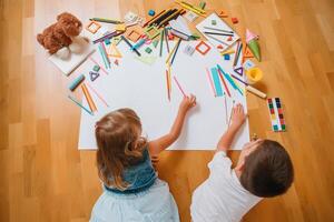 crianças desenhando em chão em papel. pré escola Garoto e menina jogar em chão com educacional brinquedos - blocos, trem, Ferrovia, avião. brinquedos para pré escola e Jardim da infância. crianças às casa ou creche. topo visualizar. foto