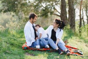 feliz jovem família gastos Tempo ao ar livre em uma verão dia ter Diversão às lindo parque dentro natureza enquanto sentado em a verde grama. feliz família. foto