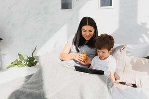 lindo sorridente mãe e filho jogando com digital tábua deitado em cama. foto