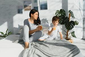 mãe com filho dentro pijamas bebendo suco dentro a manhã dentro cama. saudável comendo conceito foto