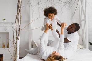 animado multirracial jovem família com pequeno menina criança ter diversão, muito feliz feliz. internacional mãe e Papai jogar com pequeno garota, apreciar final de semana às casa juntos foto