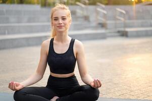 ioga e meditação dentro moderno cidade caucasiano mulher relaxar lótus posição sentado ioga esteira ao ar livre verão parque em concreto chão foto