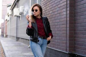 lindo jovem elegantemente vestido caucasiano menina fuma cigarro em a rua. foto