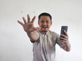 adulto ásia homem sorridente e acenando dele mão enquanto segurando Móvel telefone foto