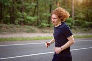 caucasiano jovem encaracolado homem atleta corre ensolarado verão dia em asfalto estrada dentro a floresta. foto