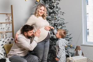 Natal família felicidade retrato do pai, grávida mãe e pequeno filho sentado poltrona às casa perto Natal árvore abraço sorrir foto