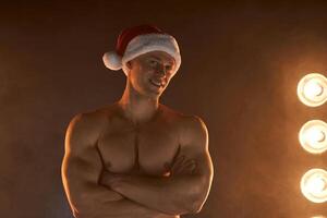 retrato do muscular homem vestindo Natal santa chapéu, guardada mãos em esfumaçado fundo foto