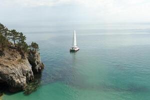 panorâmico Visão do pacífico oceano azul transparente água, uma branco esporte moderno luxo iate vela barco flutuando e uma verde costa com floresta, colinas. feriado de a água e vela corrida. foto