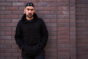 bonito jovem com a barba por fazer homem Preto moletom com capuz e bandana carrinhos quintal perto tijolo paredes verão dia. foto