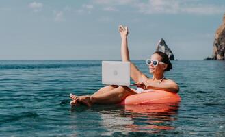 mulher trabalhador autonomo trabalho em computador portátil natação dentro mar em Rosa inflável anel. feliz turista dentro oculos de sol flutuando em inflável rosquinha e trabalhando em computador portátil computador dentro calma oceano. controlo remoto trabalhando qualquer lugar foto