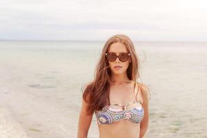 retrato do uma lindo sexy jovem mulher em a praia, bronzeada radiante pele, grandes saudável cabelo, bikini foto