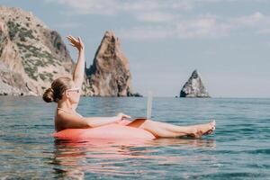 mulher trabalhador autonomo trabalho em computador portátil natação dentro mar em Rosa inflável anel. feliz turista dentro oculos de sol flutuando em inflável rosquinha e trabalhando em computador portátil computador dentro calma oceano. controlo remoto trabalhando qualquer lugar foto