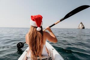 mulher dentro caiaque costas visualizar. feliz jovem mulher dentro santa chapéu flutuando dentro caiaque em calma mar. verão feriado período de férias e alegre fêmea pessoas relaxante tendo Diversão em a barco. foto