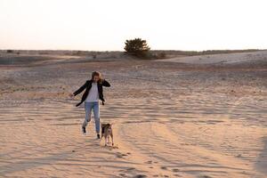 uma jovem caucasiano menina em algum lugar dentro uma couro Jaqueta e azul jeans corre ao longo a arenoso de praia com dela beagle cachorro foto