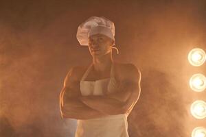 retrato do muscular chefe de cozinha vestindo branco avental e chefe de cozinha chapéu, posando com guardada braços em esfumaçado fundo foto