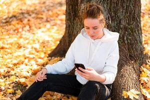 mulher dentro uma branco suéter com uma de capuz senta em terra dentro a parque perto a árvore e segurando uma celular dentro dela mãos foto