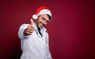 jovem bonito médico dentro branco uniforme e santa claus chapéu em pé dentro estúdio em vermelho fundo sorrir e dedo dentro Câmera foto