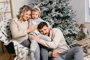 Natal família felicidade retrato do pai, grávida mãe e pequeno filho sentado poltrona às casa perto Natal árvore abraço sorrir foto