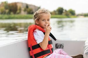 pequeno menina vestido vida colete passeio prazer barco em cidade rio ensolarado verão dia caucasiano fêmea criança natação em barco foto