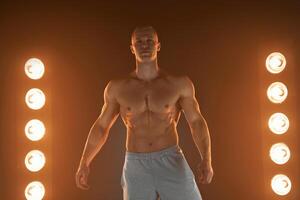 ativo estilo de vida. profissional fisiculturista mostrando perfeito muscular corpo e sorridente às Câmera, lâmpadas iluminação em fundo foto