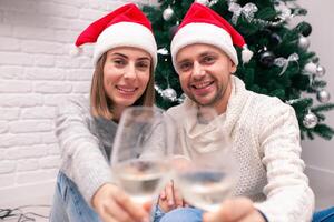 jovem feliz casal dentro Natal chapéus perto uma Natal árvore e segurando óculos do vinho. Novo ano celebração foto
