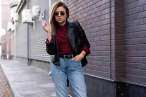 lindo jovem elegantemente vestido caucasiano menina fuma cigarro em a rua. foto