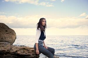 o negócio mulher em terno colete e branco camisa em pé perto grande pedra em a mar custo foto