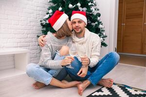 jovem feliz casal dentro Natal chapéus perto uma Natal árvore se beijando, segurando óculos do vinho. Novo ano celebração foto