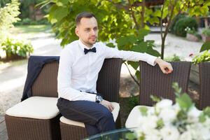 uma bonito jovem o negócio homem dentro uma branco camisa e gravata uma borboleta às uma o negócio jantar espera para dele colegas sozinho às uma verão restaurante mesa foto