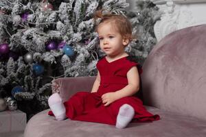 lindo fofa pequeno menina sentado em a sofá perto a Novo ano árvore. feriado humor, infantil espontaneidade e doce atmosfera foto