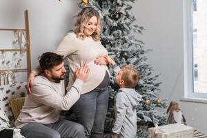 Natal família felicidade retrato do pai, grávida mãe e pequeno filho sentado poltrona às casa perto Natal árvore abraço sorrir foto