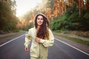 lindo caucasiano jovem menina atleta corre ensolarado verão dia em asfalto estrada dentro a floresta. foto