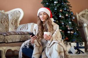 Natal. mulher vestido branco suéter santa chapéu em a chão perto Natal árvore com smartpone e copo do café foto