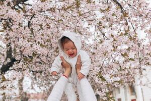 retrato do feliz alegre criança dentro branco roupas sobre árvore flores Flor fundo. família jogando juntos fora. mãe alegremente aguarde pequeno filha foto